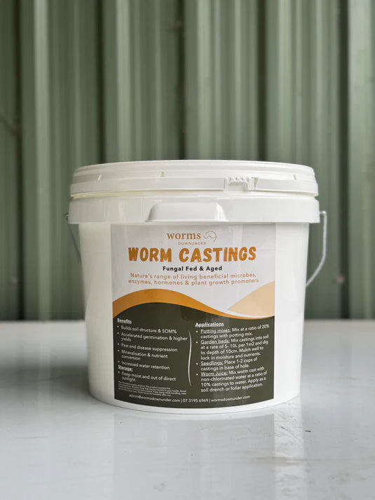 A white plastic bucket of "Worm Castings – Microbe-Rich Organic Soil Booster" with a beige and brown label sits on a white surface, promoting vibrant, healthy plants perfect for wildlife gardens, set against a corrugated green metal background.
