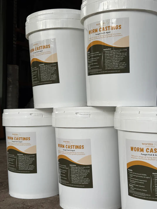Five large white plastic buckets with sealed lids, labeled “Worm Castings – Microbe-Rich Organic Soil Booster,” are stacked on a concrete floor. The attractive green and beige labels showcase the organic blend’s ability to enrich soil, boost plant growth, and attract beneficial wildlife to your garden.
